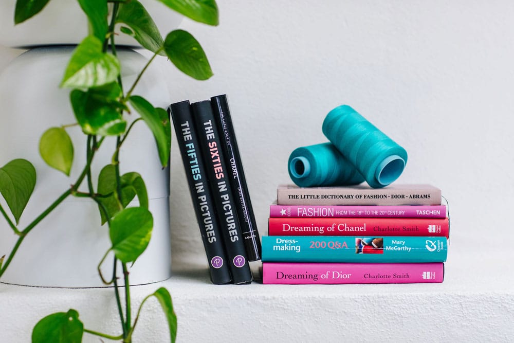 A neat row of fashion-themed books between a plant and blue dumbbells on a white contact shelf. A neat row of fashion-themed books between a plant and blue dumbbells on a white contact shelf.