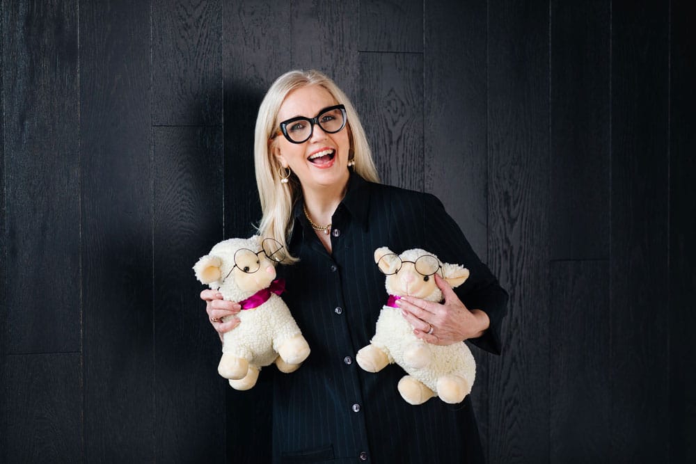 A smiling woman with glasses, wearing a black blazer designed by a sustainable fashion educator, holds two plush sheep toys against a dark wooden background. A smiling woman with glasses, wearing a black blazer designed by a sustainable fashion educator, holds two plush sheep toys against a dark wooden background.