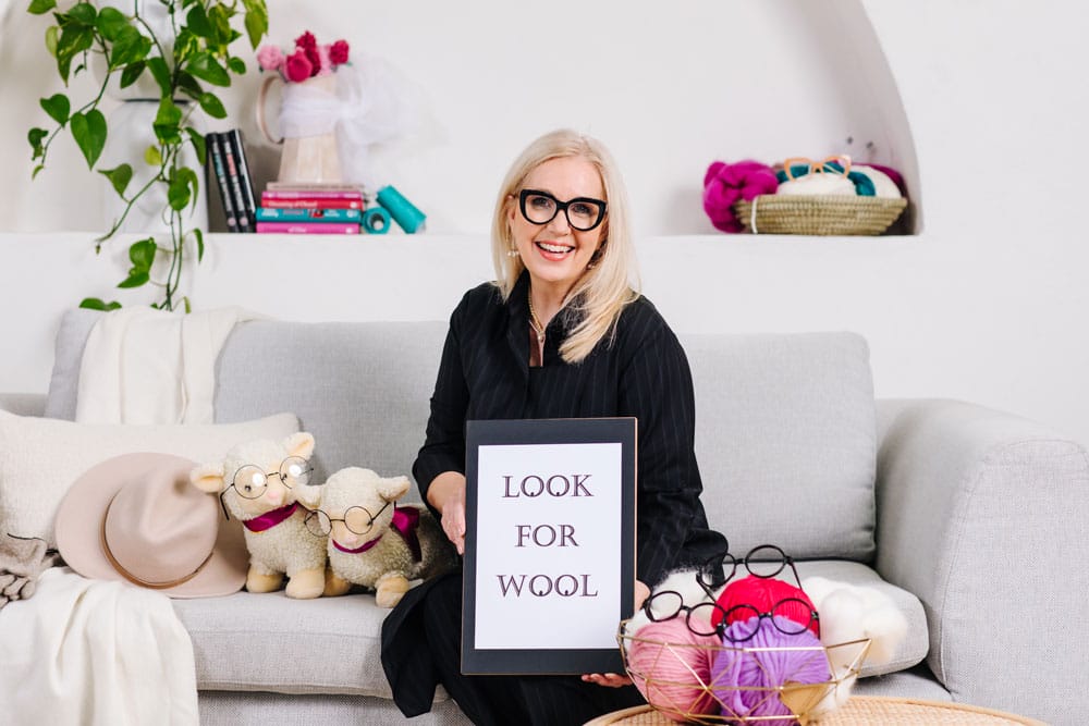 sue-maree-dazzle-image-consulting A smiling woman with glasses showing colorful fabrics against a dark wooden background.